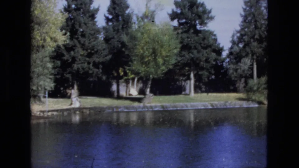 1974 Colorado Springs 8mm frame showing a calm pond bordered by a low stone wall, with evergreen and deciduous trees across the water.
