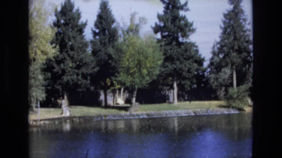 1974 Colorado Springs 8mm frame showing a calm pond, grassy bank, and tall evergreen trees with soft home-movie color and dark film edges.
