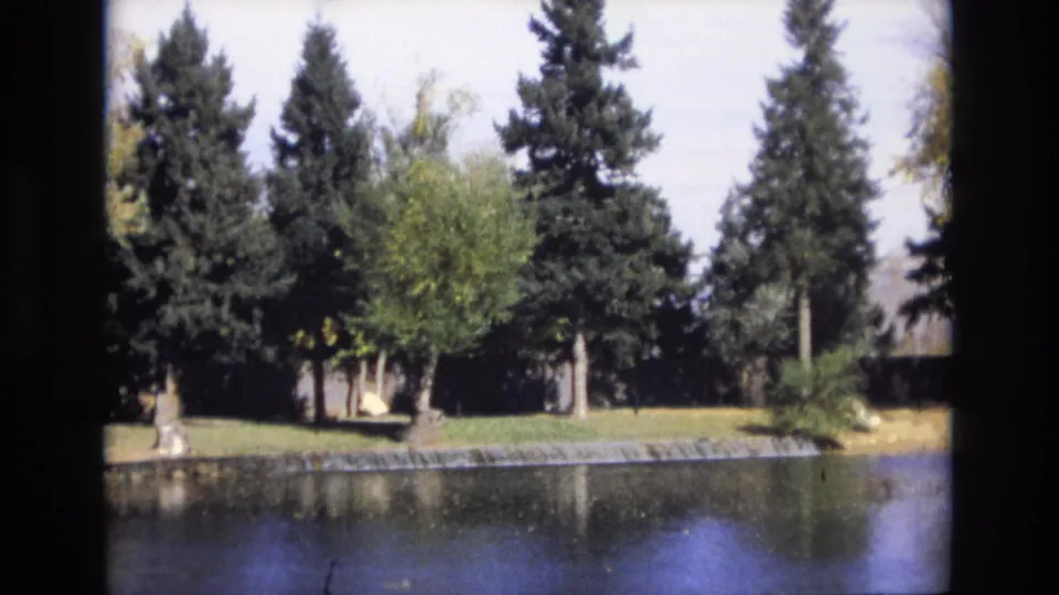 1974 Colorado Springs frame showing a quiet lake, reflected evergreens, a grassy shore, a low retaining edge, and a small pale background structure partly screened by foliage.