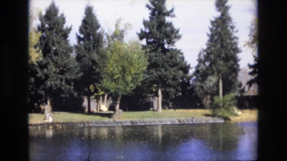 1974 Colorado Springs 8mm frame showing a calm pond, a low waterfall or spillway, tall evergreens, and mixed trees along the opposite bank.