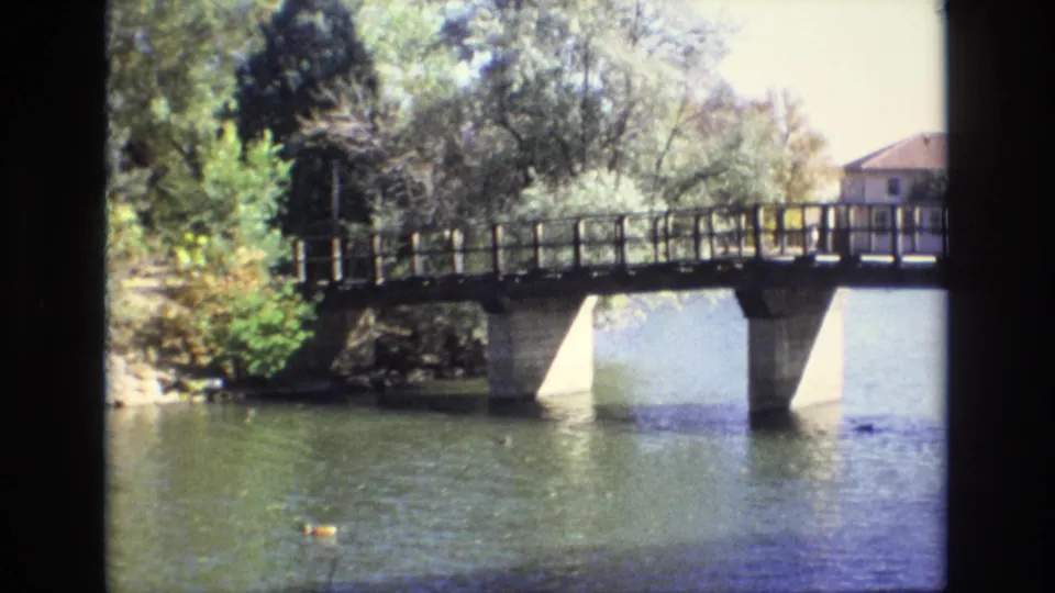 1974 Colorado Springs 8mm frame showing a pedestrian bridge over a pond or lake, with trees, shrubs, ducks on the water, and a distant building partly visible.