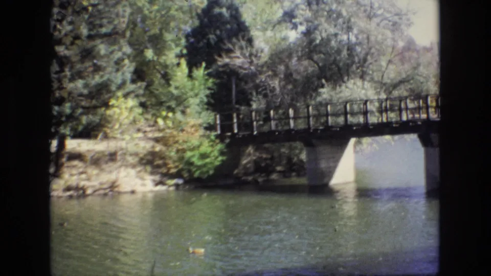 1974 Colorado Springs 8mm frame of a bridge spanning a pond or lake, with ducks on the water, dense trees, shoreline rocks, and a partially obscured background building.