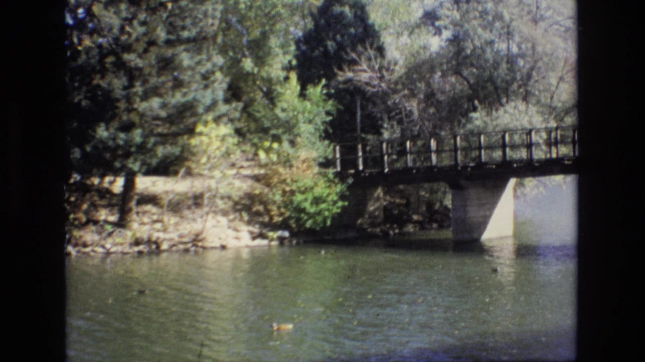 1974 Colorado Springs 8mm frame showing a calm pond with ducks in the foreground, a small wooden bridge, trees, and a shaded rocky bank.