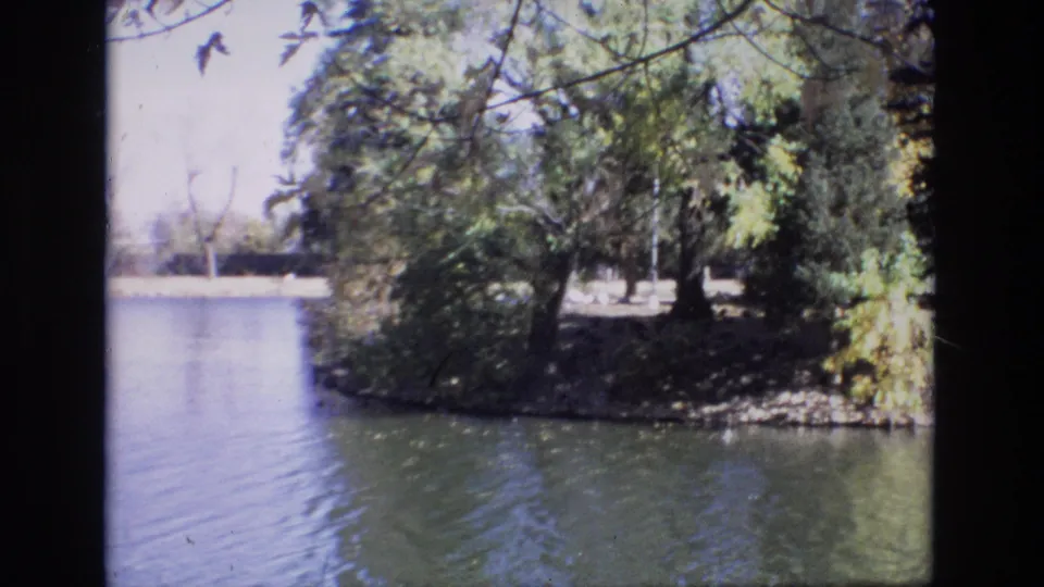 1974 Colorado Springs home-movie frame showing a calm lake, tree-lined shore, and overhanging branches in soft 8mm color.
