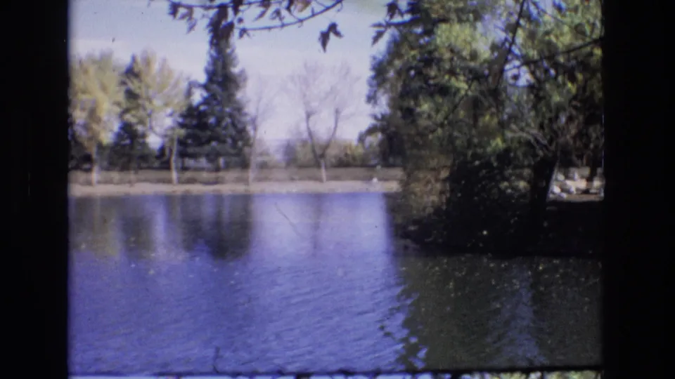 1974 Colorado Springs 8mm frame showing a broad lake view with rippled water, trees, and a shaded bank.