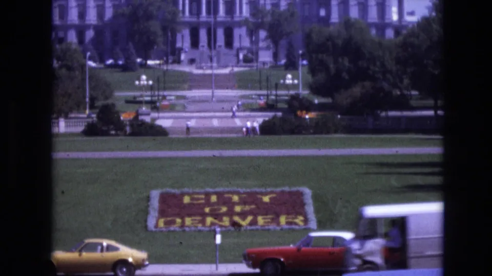1974 Denver street view showing the floral "CITY OF DENVER" sign on a grassy slope, with a yellow coupe, red car, pedestrians, trees, and a large civic-looking building beyond.
