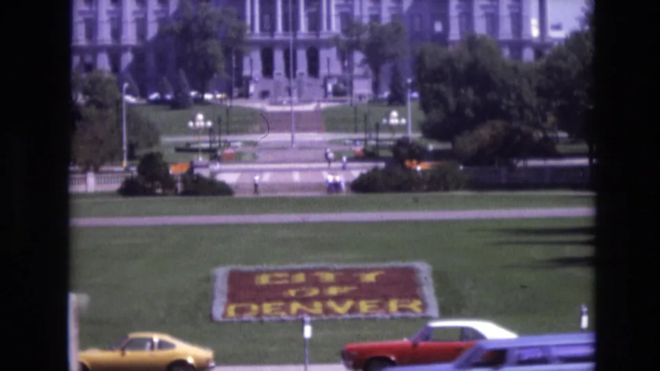 1974 Denver 8mm frame with the "CITY OF DENVER" floral display, a yellow car and red convertible passing in the foreground, pedestrians on the grounds, and a large formal building in the background.