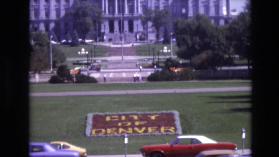 Late frame from the 1974 Denver clip showing the floral "CITY OF DENVER" sign on the lawn, with a red convertible, yellow car, pedestrians, trees, and the large background building softened by 8mm focus.