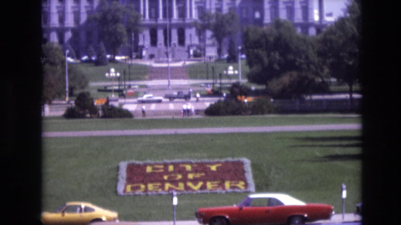 1974 Denver home-movie frame showing a floral CITY OF DENVER sign on a green lawn, a yellow coupe and red-and-white car passing, and a large formal building beyond the street.