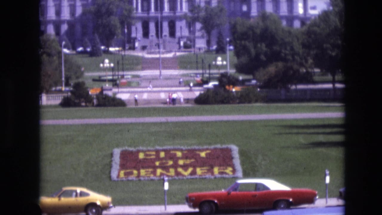 1974 Denver frame with the CITY OF DENVER floral sign centered on the lawn while a yellow coupe and red-and-white car move along the foreground street.