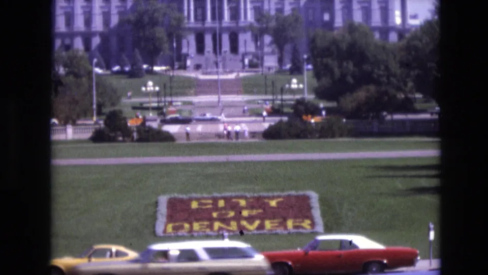 1974 Denver home-movie frame showing a white station wagon and red car crossing the foreground, with the floral CITY OF DENVER sign and civic building visible behind them.