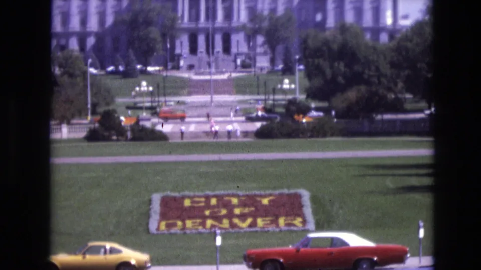 Final 1974 Denver frame with a red-and-white car and yellow coupe near the foreground street, the floral CITY OF DENVER sign on the lawn, and pedestrians near the distant building.