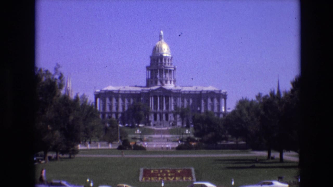 1974 8mm home-movie frame showing the Colorado State Capitol in Denver across a green lawn, with trees, parked cars, and a City of Denver sign in the foreground.