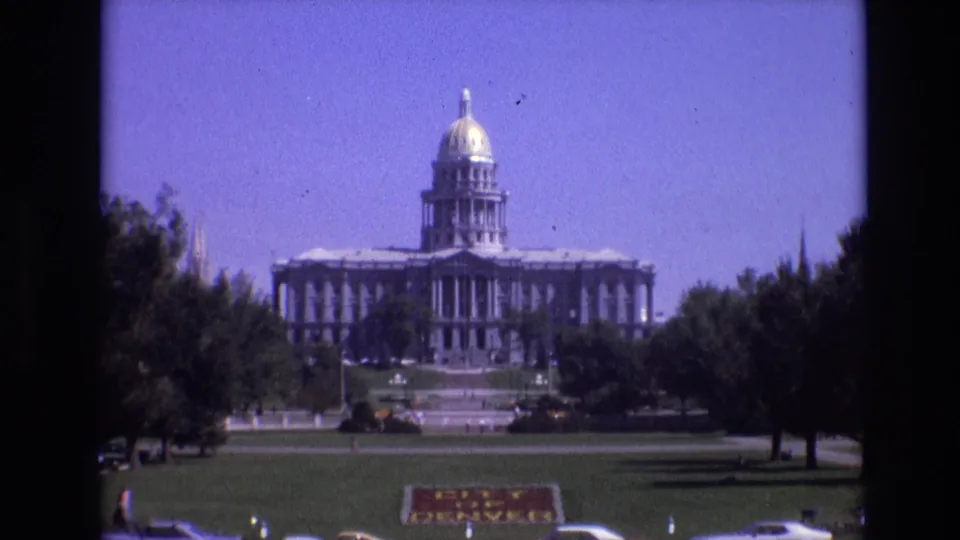 1974 8mm home-movie frame showing the Colorado State Capitol in Denver across a green lawn, with trees, parked cars, and a City of Denver sign in the foreground.