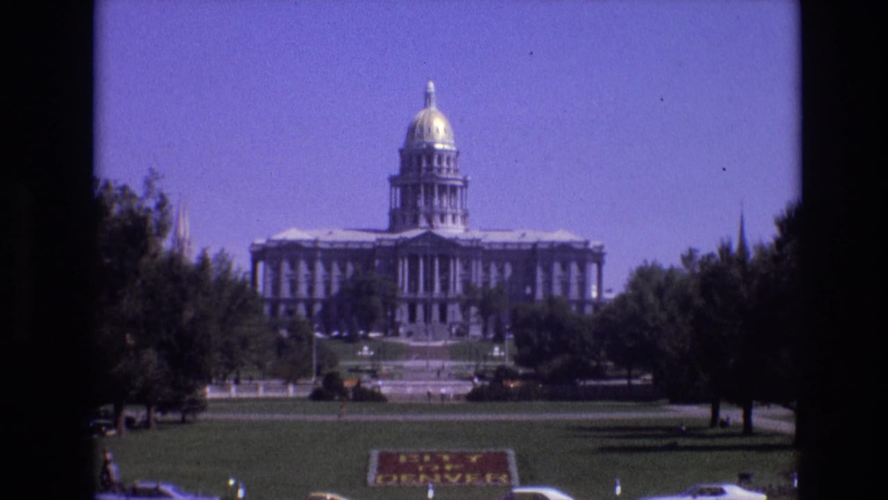 Wide capitol view framed by trees with lawn, sign, and cars in the foreground.