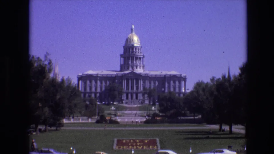 1974 Denver frame of the Colorado State Capitol with its gold dome centered beyond a lawn, trees, a City of Denver sign, and parked cars.