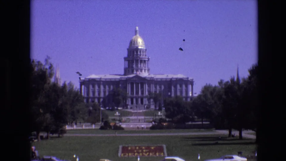 1974 8mm frame showing a closer visitor-shot view of the Colorado State Capitol in Denver, with the dome, columns, lawn, trees, parked cars, and City of Denver sign visible.