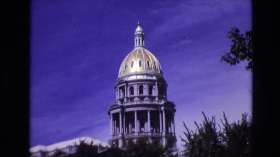 Close 1974 Denver home-movie frame of the Colorado State Capitol dome and upper colonnade, partly framed by tree branches and dark film borders.