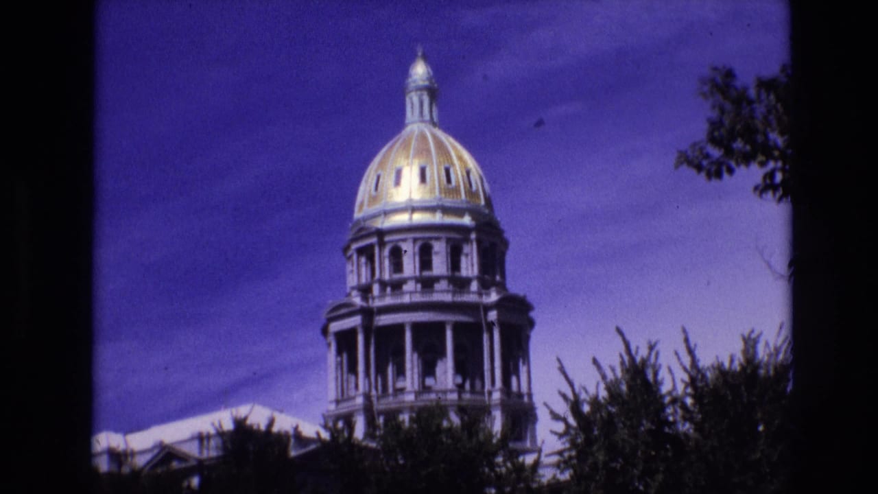 1974 Denver home-movie frame showing the gold dome and upper columns of the Colorado State Capitol above trees under a saturated blue-purple sky.