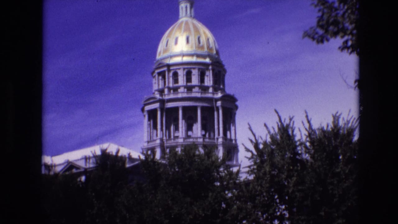 1974 Denver frame with the Colorado State Capitol dome and columned drum partly obscured by dark trees and film-frame edges.