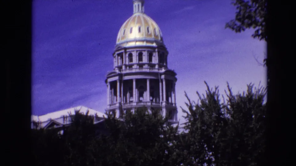 1974 Denver frame with the Colorado State Capitol dome and columned drum partly obscured by dark trees and film-frame edges.