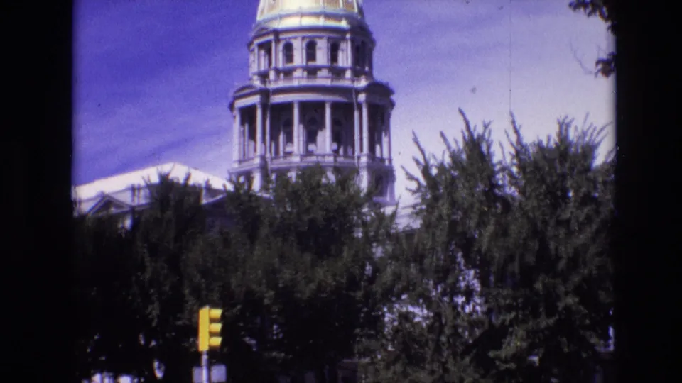 1974 Denver home-movie frame widening from the capitol dome to show dense trees and a yellow traffic light below the building.