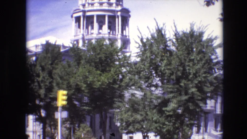 1974 Denver frame showing the capitol building partly hidden by green trees, with a yellow traffic light visible in the foreground.