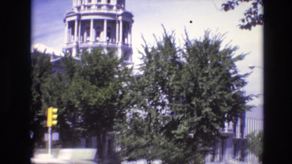 1974 Denver street view with a domed classical building partly obscured by leafy trees and a yellow traffic signal.
