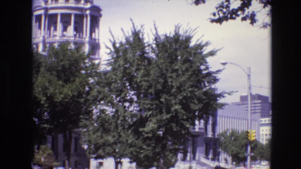 Denver street scene from 1974 showing a domed building behind trees, a curved streetlamp, yellow traffic signal, and high-rise buildings in the background.