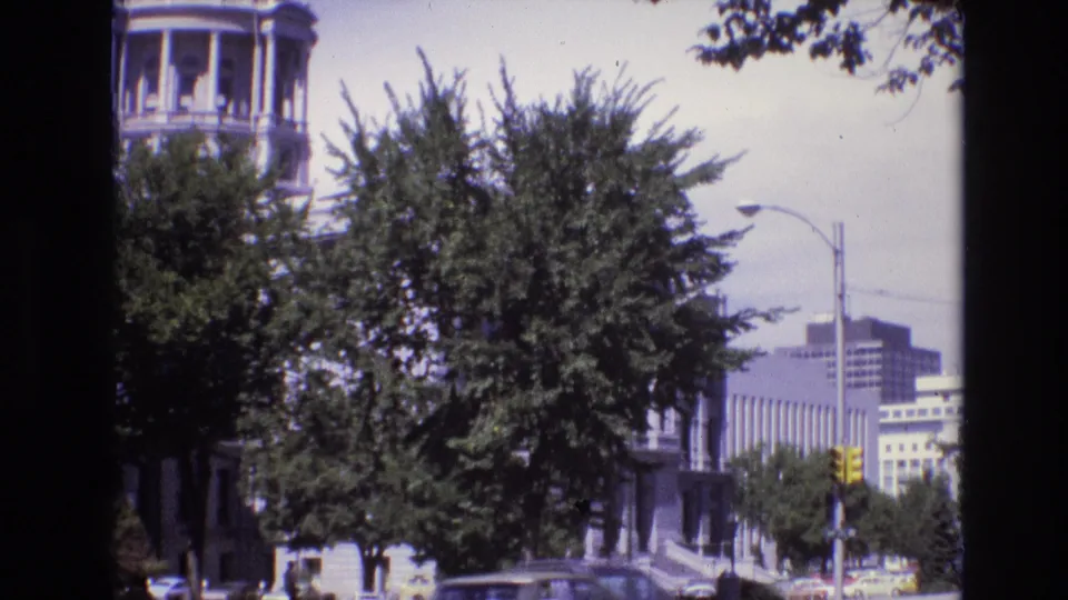 1974 Denver downtown view with trees obscuring a classical building, a yellow traffic light, background office buildings, and cars along the street.