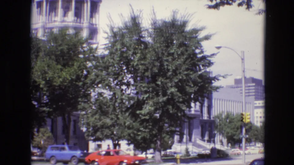 Denver street view from 1974 with a classical building and steps partly hidden by trees, yellow traffic signals, a streetlamp, and red and blue cars passing or parked nearby.
