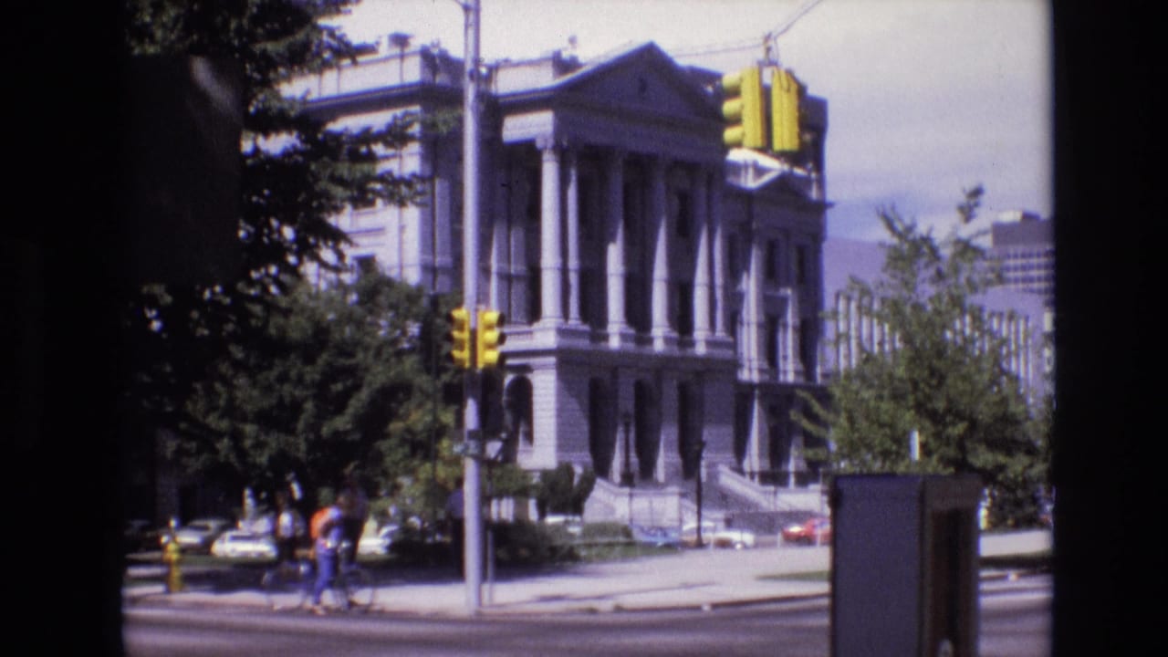 1974 Denver street view of a neoclassical civic building with columns, yellow traffic lights, cars, pedestrians, and a cyclist near the corner.