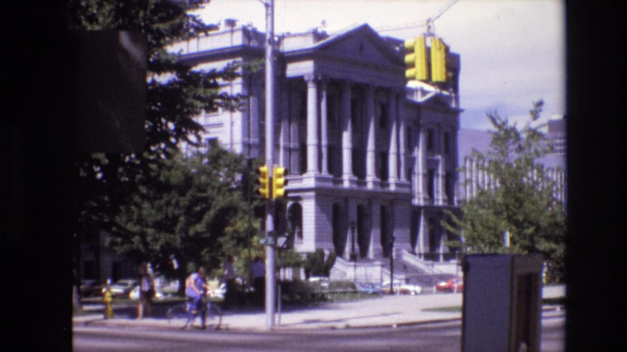 1974 Denver frame showing pedestrians and a bicycle near a traffic signal, with a large columned civic building and trees in the background.