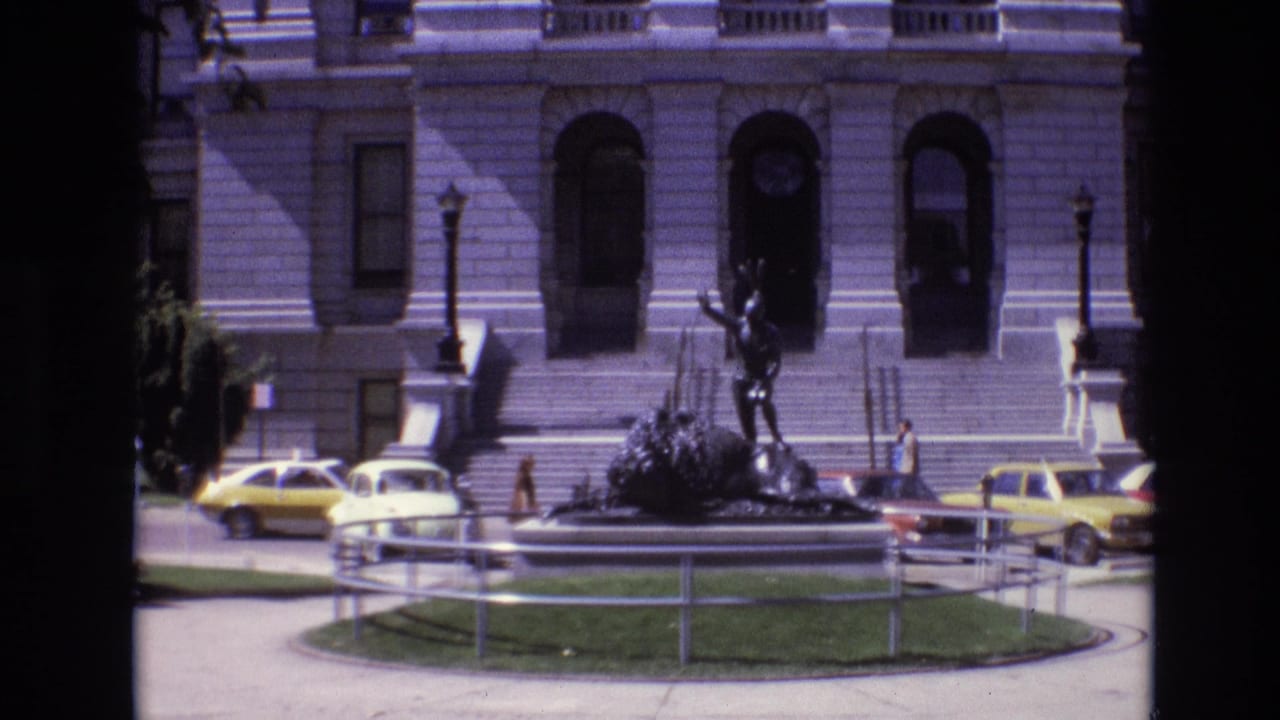 Neoclassical civic building behind a bronze statue and parked 1970s cars.
