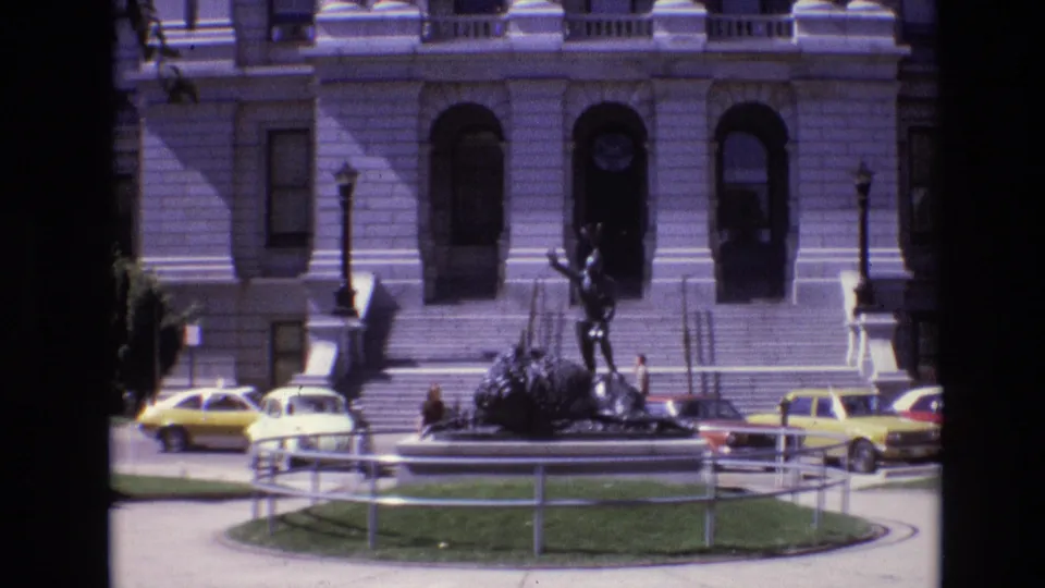 1974 Denver frame with a bronze statue centered before a grand stone government building, with steps, arched windows, railings, and period cars along the curb.