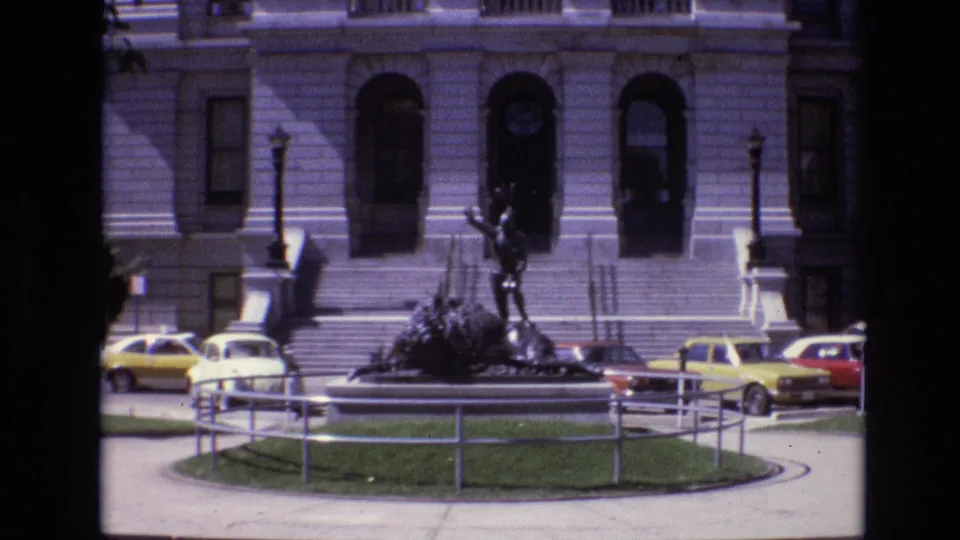 1974 Denver home-movie view of a bronze statue on a circular grass island, with railings, stone steps, arched windows, and yellow and white cars in the background.