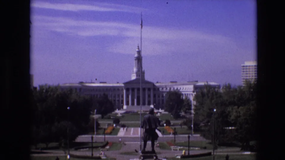 1974 Denver frame looking across a landscaped civic plaza toward a large government building with a central tower, flagpole, trees, paths, benches, and a statue in the foreground.
