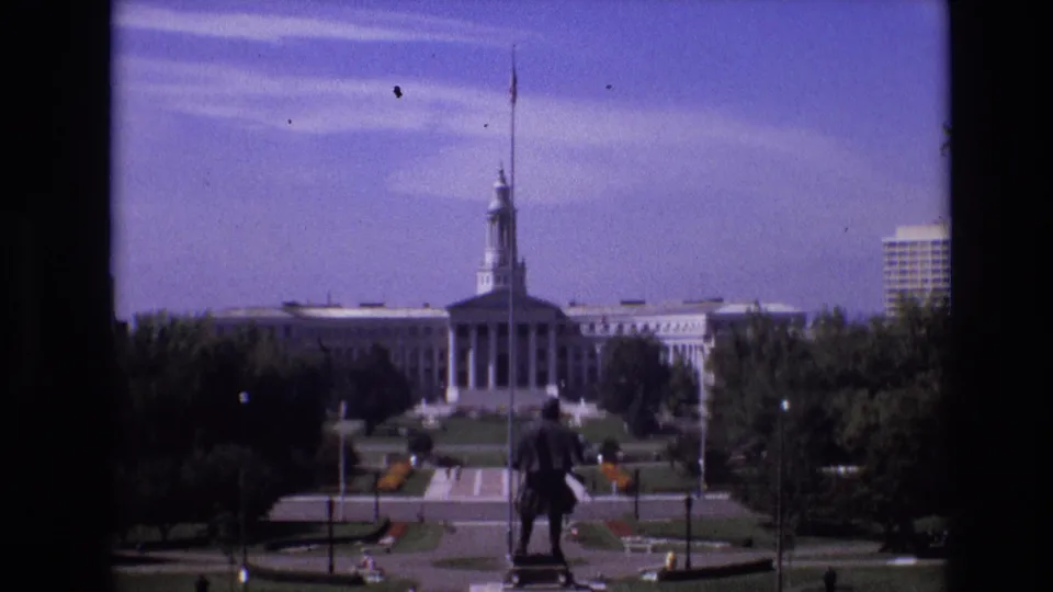 1974 Denver 8mm frame showing the Colorado State Capitol across a landscaped civic park, with a statue in the foreground and small pedestrians on the paths.
