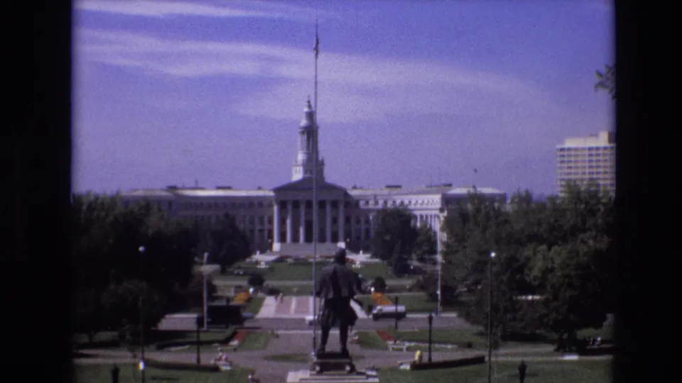 1974 Denver 8mm still showing a visitor's wide view toward the Colorado State Capitol, with the statue, landscaped park, cars, and pedestrians visible at a distance.