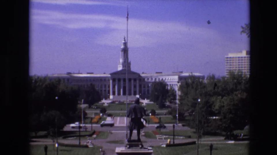 1974 Denver frame showing the Capitol across a formal green space, with a statue in the foreground, tree cover along the sides, and light traffic near the lower edge.