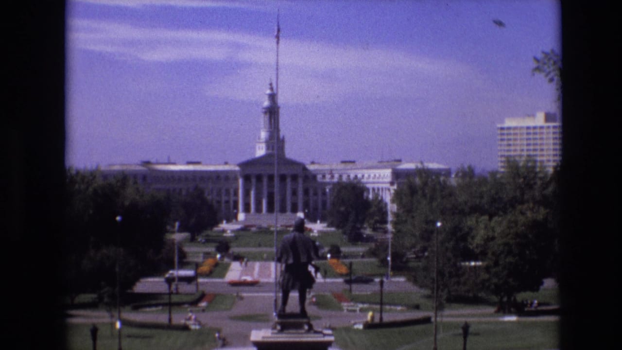 1974 Denver home-movie frame showing a distant standing monument in a landscaped park, with pedestrians, lawns, flowerbeds, roads, and a large classical civic building beyond.