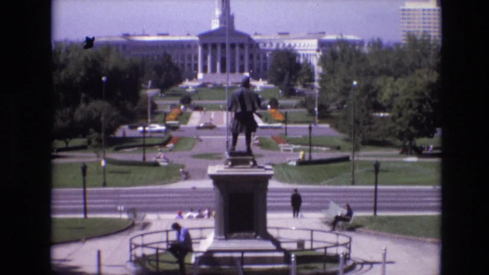 1974 Denver park view with a standing statue on a pedestal in the foreground, seated and walking visitors nearby, and a large civic building framed by trees in the background.