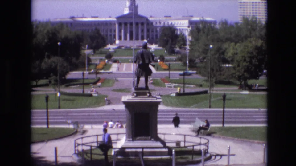1974 Denver 8mm frame showing the monument centered above a road and park paths, with seated visitors, small groups on the lawn, trees, and a towered civic building behind.