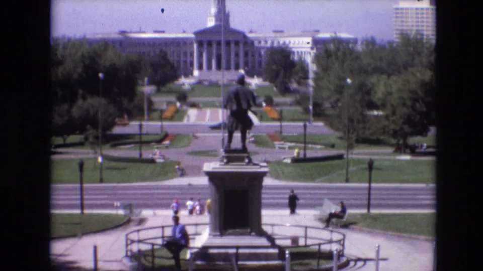 1974 Denver home-movie frame with the statue and pedestal centered against lawns, paths, a few pedestrians, flowerbeds, and the large civic building beyond the park.