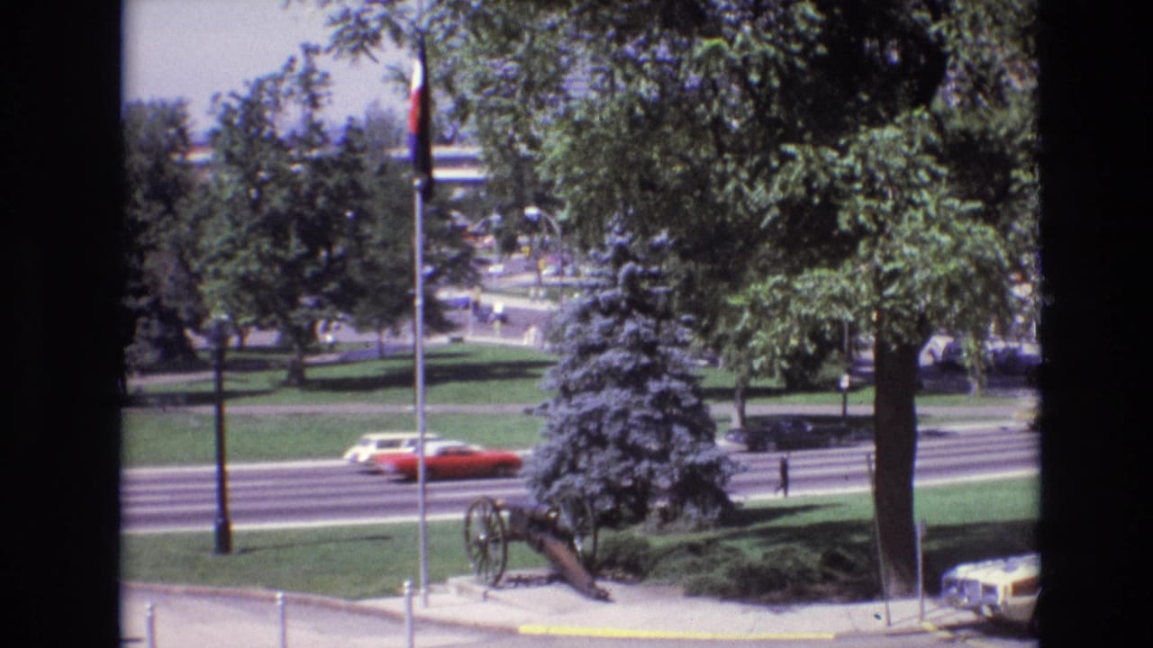 1974 Denver street scene with a red car moving along a multi-lane road beside a landscaped park, flagpole, trees, and a historic cannon display.