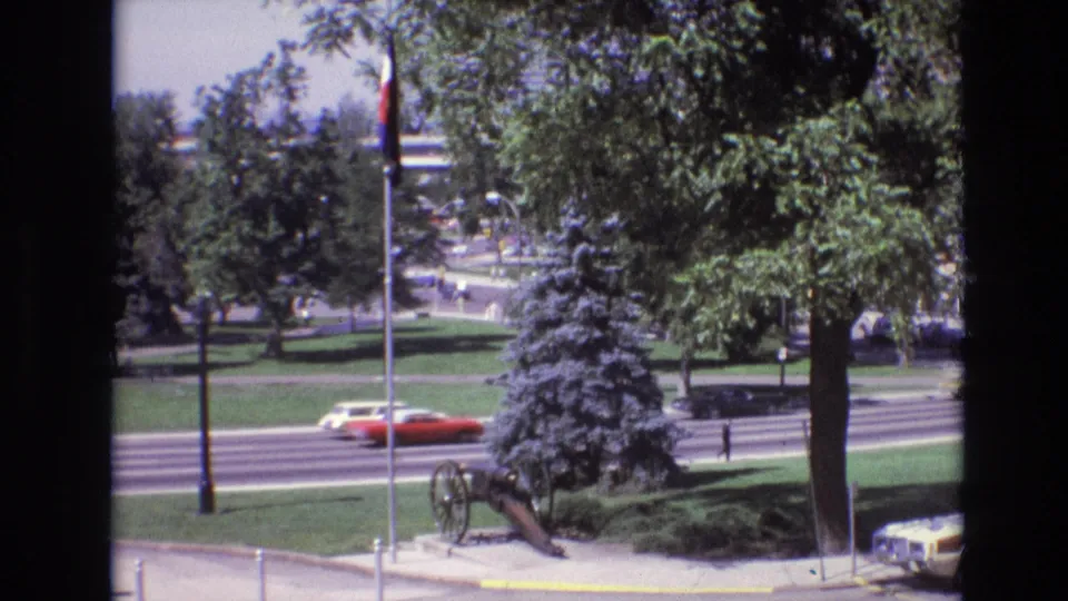 1974 Denver street scene with a red car moving along a multi-lane road beside a landscaped park, flagpole, trees, and a historic cannon display.
