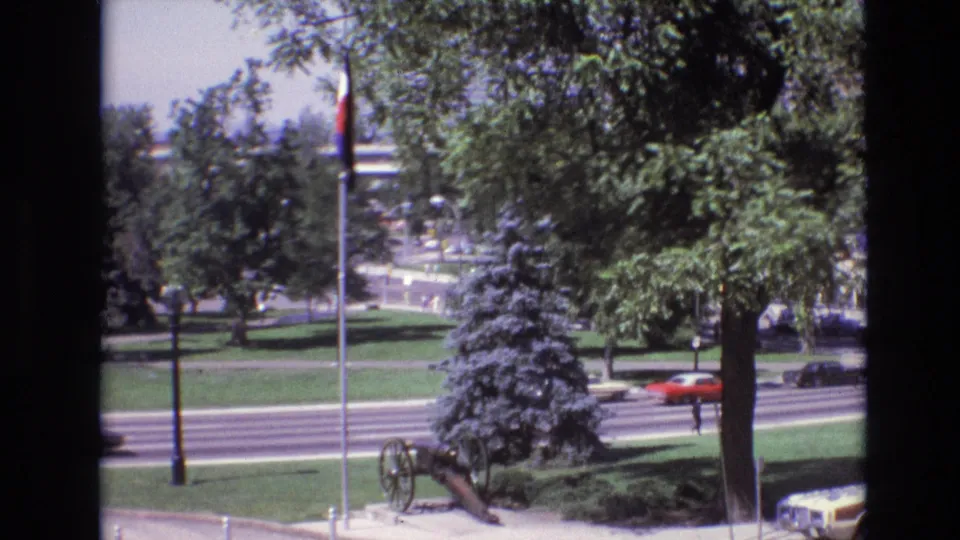 1974 Denver home-movie frame showing a red car on the roadway, a pedestrian-sized figure near the street, and a civic park with trees, flagpole, and cannon display.
