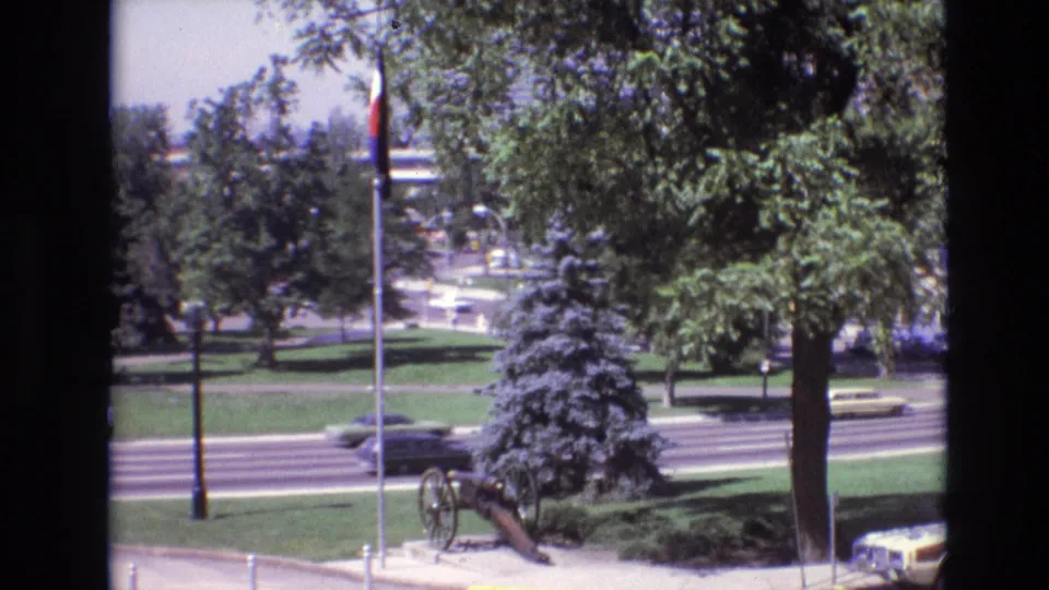 1974 Denver 8mm frame with motion-blurred traffic passing a civic park, where a flagpole, cannon display, lawns, trees, and distant buildings remain visible.