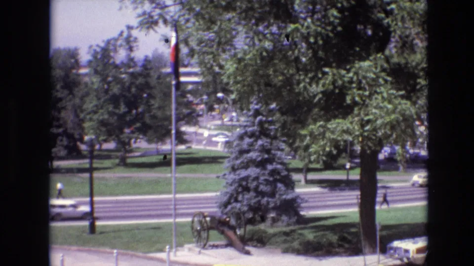 1974 Denver home-movie frame showing a historic cannon in a landscaped park near a flagpole, with trees, lawns, and traffic beyond.
