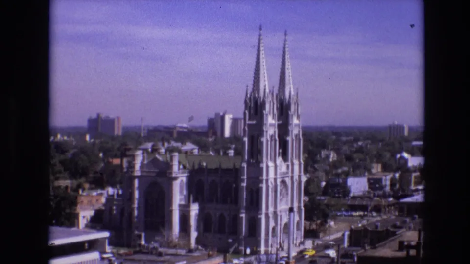 1974 Denver skyline frame centered on a Gothic-style cathedral with tall twin spires, neighboring buildings, streets, and distant towers.